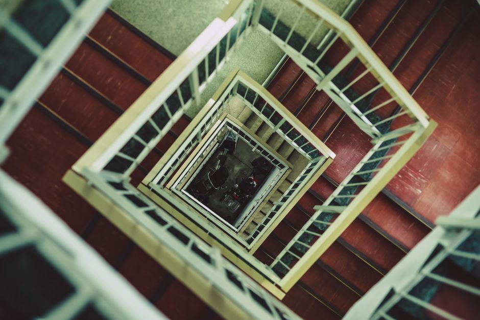A top-down view of a multi-story staircase inside a building, with a spiral design creating a square pattern as the staircase descends through several levels. The treads are made of dark wood, while the railing and balustrades are painted in a pale color, possibly cream or white. The central opening shows areas of the staircase at different heights, with the bottom visible where items such as black cardboard boxes and plastic wrapping materials are being prepared for moving. The environment is well-lit with natural or artificial light, highlighting the clean and organized appearance typical of professional house removal and furniture transport services. The image captures the process of moving items through a staircase during a home relocation, with the staircase providing a challenging access point that companies like Man with Van Holloway assist with during the loading process.