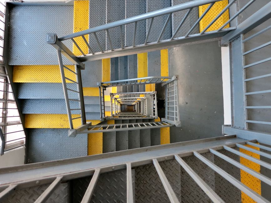 A view looking directly down a narrow, multi-level staircase in a residential building during a home relocation process, with metal railings on either side and non-slip yellow and gray treads on each flight of stairs. The staircase is enclosed within the property, with natural light filtering through windows visible on the left. The steps are supported by a sturdy metal framework, and the photograph captures the repetitive pattern of the descending staircase as it extends deeper into the building, illustrating the logistical challenge of moving large furniture and packed boxes through confined stairwells. The clean, structured environment indicates an organized moving process facilitated by Man with Van Holloway, who specialise in removals and furniture transport, ensuring safe and efficient loading, packing, and navigation through restricted staircases during house removal projects.