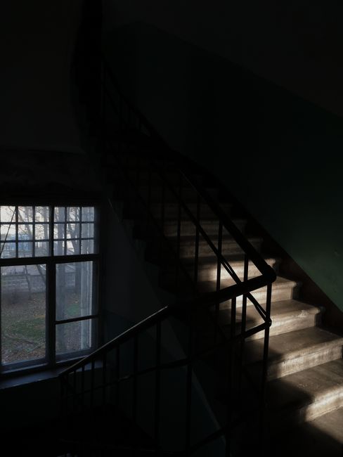 A top-down view of a multi-story staircase inside a building, with a spiral design creating a square pattern as the staircase descends through several levels. The treads are made of dark wood, while the railing and balustrades are painted in a pale color, possibly cream or white. The central opening shows areas of the staircase at different heights, with the bottom visible where items such as black cardboard boxes and plastic wrapping materials are being prepared for moving. The environment is well-lit with natural or artificial light, highlighting the clean and organized appearance typical of professional house removal and furniture transport services. The image captures the process of moving items through a staircase during a home relocation, with the staircase providing a challenging access point that companies like Man with Van Holloway assist with during the loading process.