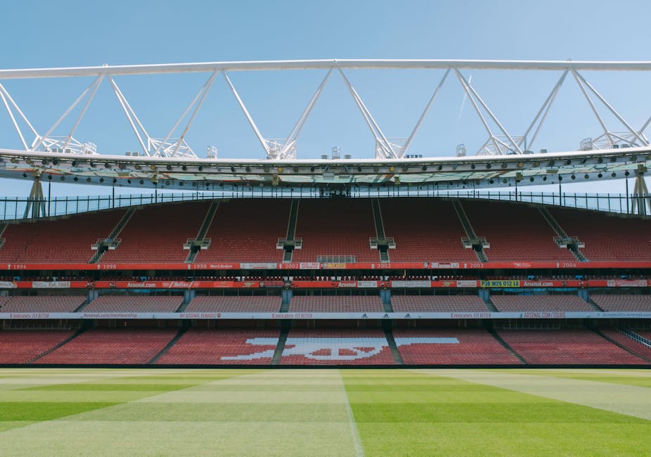 Interior view of the Emirates Stadium during daytime, showing the empty red tiered seating sections with rows of seats, some supporting structures and railings, and the bright green playing field with visible grass patterns in the foreground. The sky is clear and blue, and parts of the stadium’s roof structural framework can be seen at the top of the image. This scene illustrates the stadium's interior, possibly during a matchday or event preparation, with no people present. The image is related to house removal services in the HOLLOWAY area, captured in a professional context to suggest logistical planning for large-scale relocation or equipment transportation for sports venues, with Man with Van Holloway’s expertise in moving large items or equipment associated with such venues evident in the surroundings.