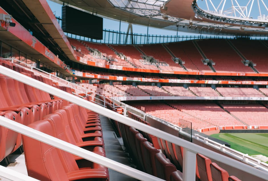 A view of the interior of the Emirates Stadium, showing multiple rows of red stadium seats arranged along a curved section of the seating area, with some seats in the foreground and additional seating levels visible in the background. The structure is illuminated by natural daylight, and the field can be seen in the distance at the bottom right corner. The image captures the spaciousness and tiered arrangement typical of the stadium's seating layout, with the roof and supporting beams overhead. This setting relates to home relocation or venue transfer services by Man with Van Holloway, as part of their house removals and moving logistics for stadium or convention centre moves, emphasizing their ability to handle large-scale furniture and equipment transport in such environments.