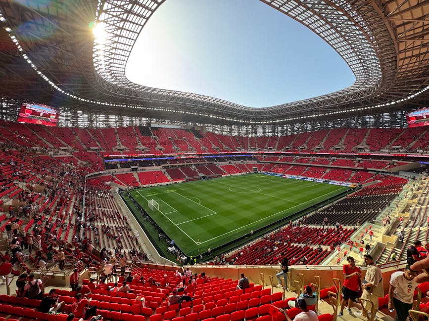 Interior view of the Emirates Stadium during daytime, showing the empty red tiered seating sections with rows of seats, some supporting structures and railings, and the bright green playing field with visible grass patterns in the foreground. The sky is clear and blue, and parts of the stadium’s roof structural framework can be seen at the top of the image. This scene illustrates the stadium's interior, possibly during a matchday or event preparation, with no people present. The image is related to house removal services in the HOLLOWAY area, captured in a professional context to suggest logistical planning for large-scale relocation or equipment transportation for sports venues, with Man with Van Holloway’s expertise in moving large items or equipment associated with such venues evident in the surroundings.