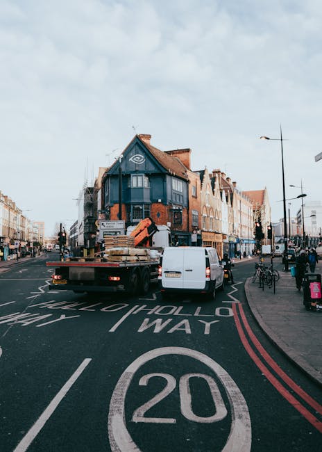 A white flatbed truck belonging to Man with Van Holloway is parked on a busy urban street in Islington, N7, near a restaurant or shop-fronts. The truck is loaded with wooden furniture, packaging materials, and cardboard boxes, some secured with straps. The vehicle is positioned across part of a lane designated for vehicle movement, with red double lines on the curb indicating parking restrictions. In the background, there are multiple multi-storey buildings with brick and tile exteriors, and several pedestrians and cyclists are visible on the sidewalk, which features lampposts and street signs. The scene is illuminated by natural daylight with a partly cloudy sky overhead. This setup illustrates the loading process involved in home relocation and furniture transport, typical of professional removals services like those offered by Man with Van Holloway, supporting efficient packing and moving activities in connection with house removals and local moving permits for N7 moves.
