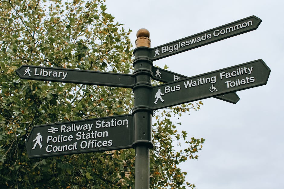 A black multi-directional street signpost featuring white text and icons, situated outdoors against a backdrop of green leafy trees and a cloudy sky. The signpost provides directions to various local landmarks and facilities, including 'Biggleswade Common', 'Library', 'Railway Station', 'Police Station', 'Council Offices', 'Bus Waiting Facility', and 'Toilets'. It has arrow-shaped signs pointing in different directions, with icons indicating pedestrian access, a train symbol for the railway station, and accessibility features for the toilets. The scene reflects a typical urban or suburban environment with clear signage for residents and visitors. Occasionally, [COMPANY_NAME] services, such as house removals and furniture transport, involve navigating such signage during home relocation or moving logistics in areas like N7, associated with [PAGE_TITLE], ensuring smooth access for loading or unloading furniture and boxes.