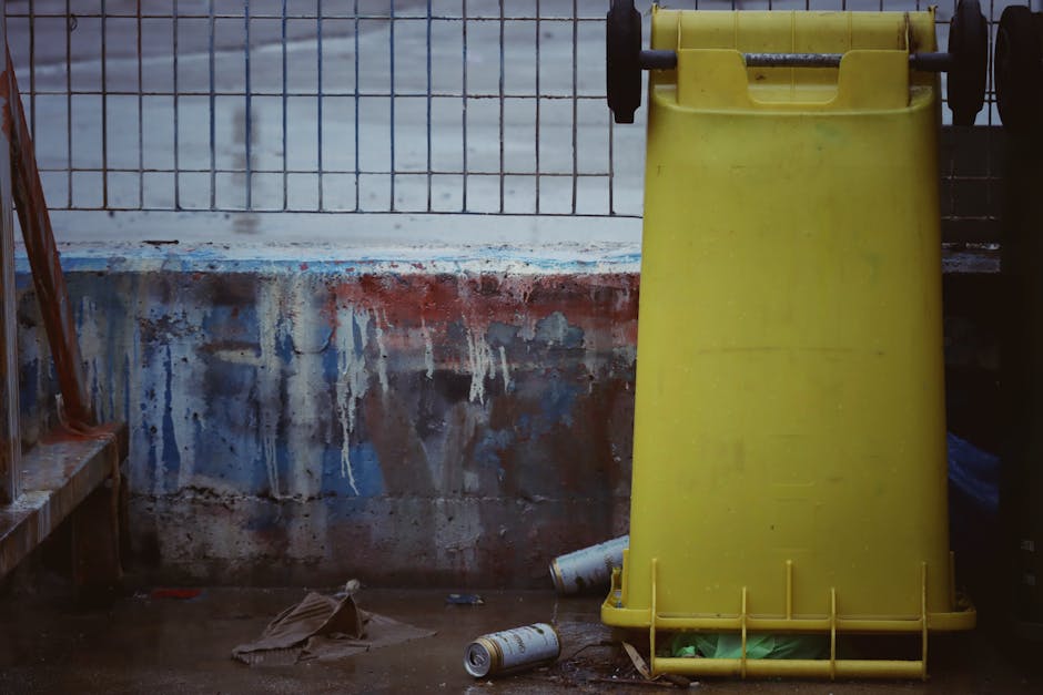 A black wheelie bin labeled 'St. John's' positioned on a sidewalk at night, with its lid open revealing various waste items including cardboard boxes, plastic packaging, and paper. The bin is located 
