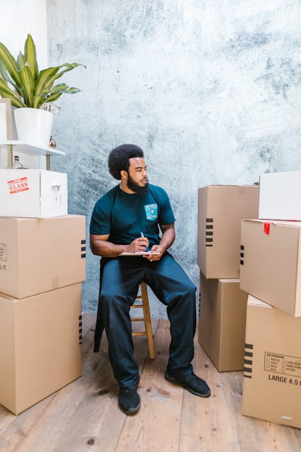 A man with dark hair and a beard, wearing a dark blue T-shirt with a logo and dark trousers, is seated on a wooden stool inside a room with a light blue, textured wall. He is holding a pen and a notep