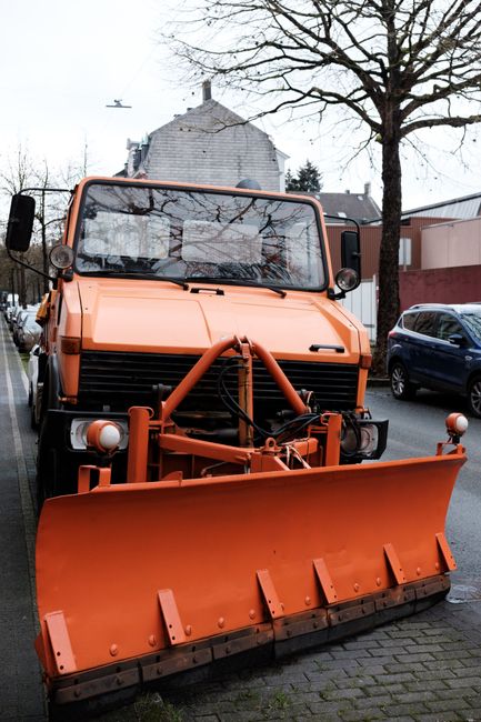 A large orange snow plough attachment is mounted to the front of a heavy-duty vehicle parked on a street. The vehicle, which appears to be used for property maintenance or removals, features a flat, rectangular windshield with a wiper, round side mirrors, and a robust exterior design. Behind the vehicle, there are residential buildings with brick walls and rooftops, along with leafless trees and a parked dark blue car to the right. The snow plough's blade is angled slightly downward and secured with metal brackets and hydraulic components. The scene is lit with natural daylight, and the setting suggests an urban environment suitable for house removals, furniture transport, or home relocation activities. The image is associated with [COMPANY_NAME], operating in the removals service category, supporting efficient loading and transport logistics for house moves.