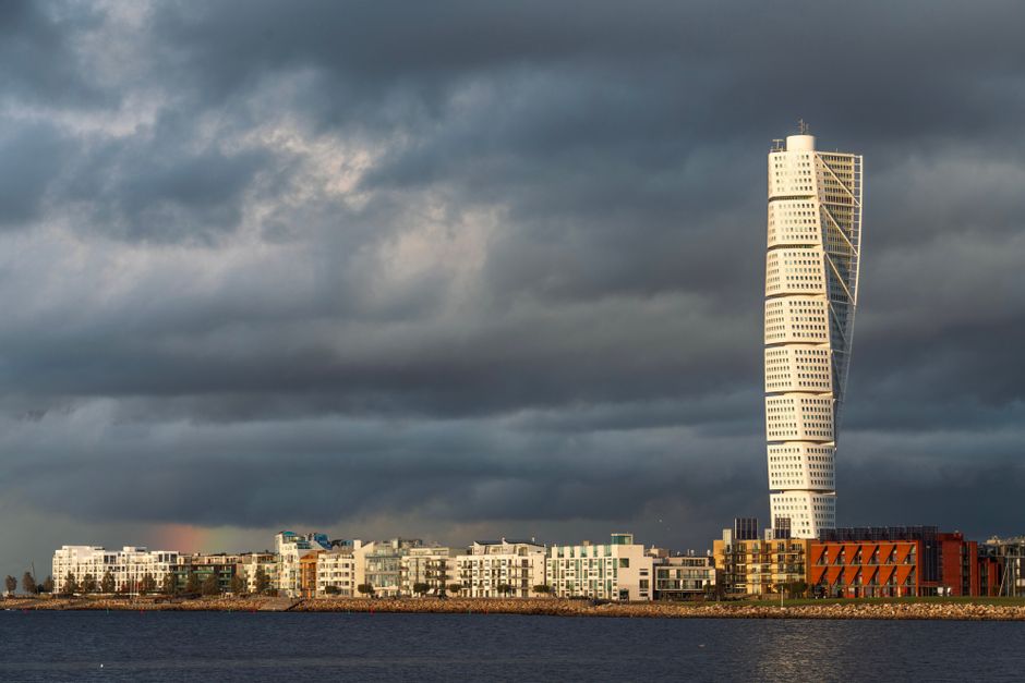 A cityscape featuring a modern, multi-storey skyscraper with a distinctive twisted design, situated behind a row of low-rise residential buildings with white facades, large windows, and balconies. The buildings are positioned along a body of water, with a low stone embankment separating them from the water. Above, dark, stormy clouds dominate the sky, casting a moody atmosphere over the scene. In the foreground, there are no visible vehicles or objects related to house removals or moving services. The image captures the urban environment possibly related to relocation activities, with a focus on the city’s architecture and weather conditions, as seen through the lens of Man with Van Holloway’s website offering removals and moving logistics in the area.