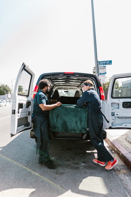 Two removal technicians from Man with Van Holloway are engaged in the home relocation process, lifting a large, heavy wardrobe wrapped in dark green protective fabric. They are placing the wardrobe into the rear of a white moving van, which has its side and back doors open, revealing a spacious interior prepared for furniture transport. The scene takes place outside on a paved street under daylight, with a street sign visible in the background. The technicians are wearing dark uniforms and safety goggles, and are utilizing proper lifting techniques to ensure the safe handling of the bulky item. Surrounding the vehicle are other parked cars and a sidewalk, indicating the clearance needed for house removals and furniture transport. This image exemplifies the packing and loading process involved in professional removals services aimed at facilitating smooth household moves, such as those highlighted in the service page for removing bulky wardrobes in Holloway.