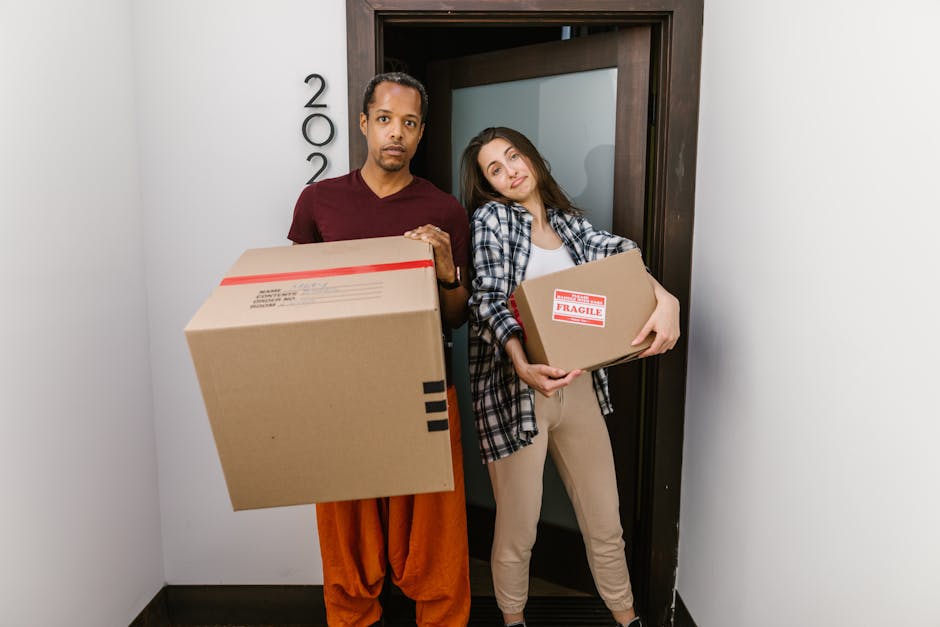 A man and a woman standing inside a doorway of a residential property, both holding cardboard boxes filled with household items; the man, dressed in a maroon T-shirt and orange pants, looks directly at the camera with a neutral expression, while the woman, wearing a checkered shirt and beige pants, leaning her head slightly, smiles faintly. The boxes, secured with red tape, are labeled with fragile handling instructions, indicating they are packed for a home relocation. The interior space has white walls, and the door frame is dark wood, suggesting a modern setting. The scene captures a moment during the packing and moving process, with the individuals preparing for furniture transport or house removals, and reflects the logistical steps involved in a professional removal service by Man with Van Holloway. The environment is well-lit, emphasizing the materials and objects being handled, such as the cardboard boxes suitable for packing household items for a furniture transport or removal service.