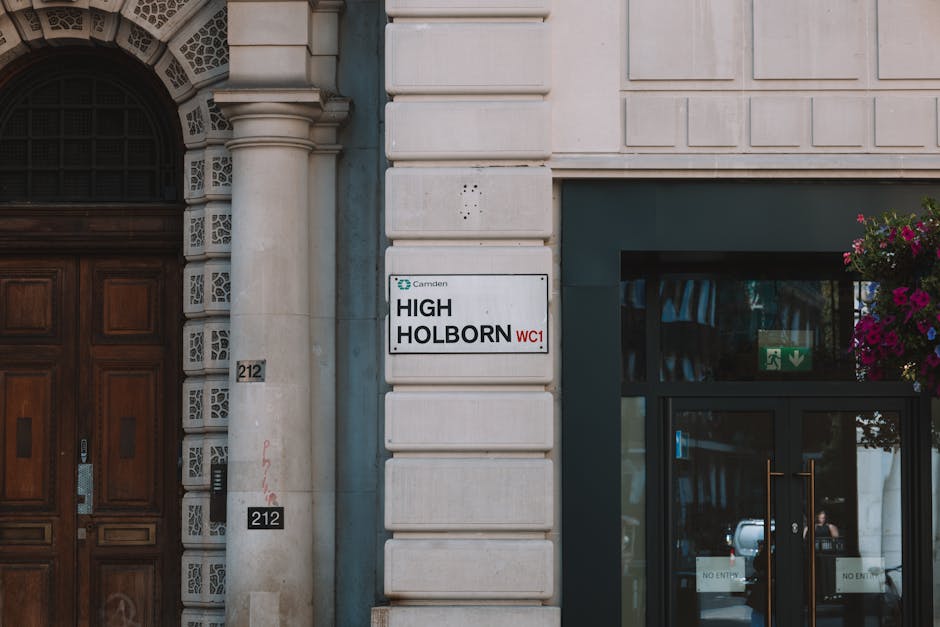 A close-up view of a building's exterior in a city environment, showing a white rectangular street sign with black and red text reading 'High Holborn WC1' attached to a light-colored stone wall. To the left is a large wooden doorway with ornate detailing, while to the right, a glass entrance door features a green emergency exit sign and a large hanging flower basket with pink and purple flowers. In the background, reflections of parked cars and street scene are visible through the glass, indicating an urban setting. The scene captures part of the building’s façade, combining architectural elements with signage typical of central London. This environment could represent a location where Man with Van Holloway offers house removals and furniture transport, emphasizing the contextual setting for relocation and moving services.