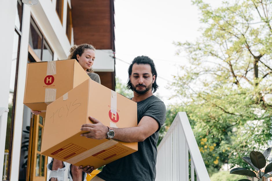 A man with dark hair and a beard, dressed in a grey t-shirt, is outside a residential property, carefully carrying two cardboard moving boxes marked with a red 'H' logo and handwritten labels, during a home relocation. Beside him, a woman with curly hair, wearing a light-colored sweater, is also holding a box, preparing to move items from the property’s doorway onto a pavement or driveway. The background features the exterior of a modern building with large windows, a white railing, and lush green trees under a clear sky, indicating daytime. The scene captures the loading process involved in packing and furniture transport, with the boxes secured with packing tape, and both individuals focusing on their task. The environment includes a partially visible van off-camera, suggesting the commencement of a move facilitated by [COMPANY_NAME], which offers removals and moving services in Holloway.